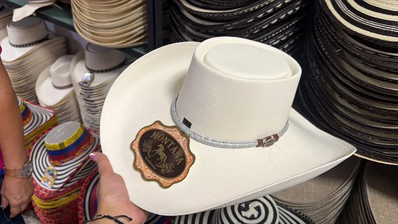 Mano sosteniendo un sombrero blanco de copa alta con cinta plateada, frente a estantes llenos de sombreros