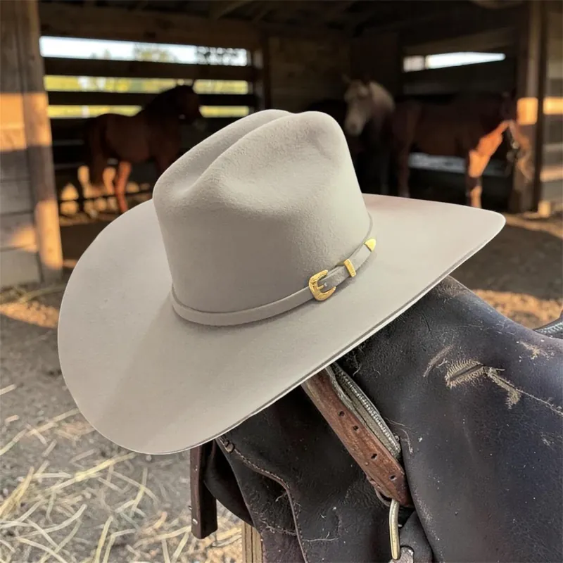 Sombrero gris de ala ancha con correa del mismo tono y hebilla dorada, sobre una silla de montar en un establo